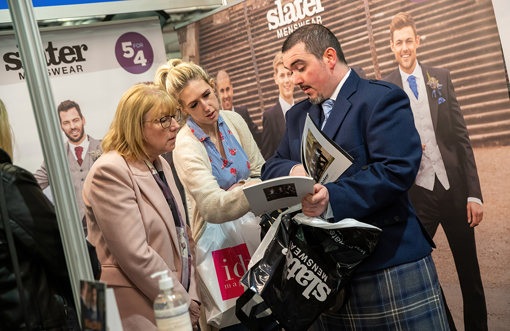 alt="Slaters mens wear member of staff showing two ladies the latest range of grooms wear in the brochure at their stand at the I Do Wedding Exhibition"